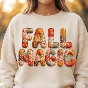 May include: A cream-colored crewneck sweatshirt featuring the text FALL MAGIC in a puffy, quilted-style font. Each letter is composed of various patchwork fabric patterns in shades of orange, yellow, brown, and sage green with floral motifs.
