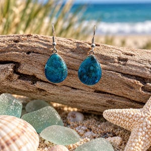 May include: A pair of teardrop-shaped earrings featuring a deep blue and turquoise mottled pattern with silver-toned metal hooks. The earrings have a smooth, glossy finish and are suspended from a textured, weathered piece of driftwood.