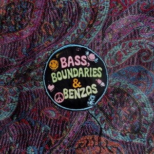 May include: A circular black embroidered patch featuring the text BASS, BOUNDARIES & BENZOS in pink, yellow, and light green. The patch includes small icons of a smiley face, a heart, a peace sign, a teddy bear, and candy, set against a paisley fabric.