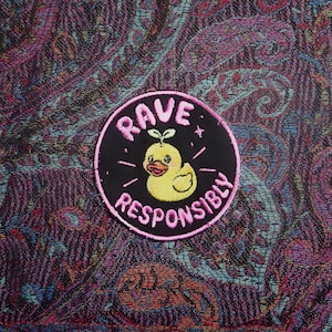 May include: A circular embroidered patch with a black background and a pink border. The center features a yellow duckling with a small green sprout on its head. The text RAVE RESPONSIBLY is embroidered in pink letters around the duckling.