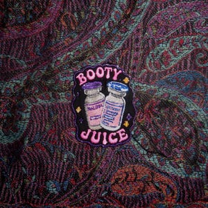 May include: An embroidered patch featuring two small glass vials with blue and purple caps, surrounded by small yellow stars. The text BOOTY JUICE is stitched in pink letters above and below the vials on a black background with a purple border.