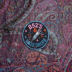 May include: A circular black embroidered patch featuring the pink text B52'S, an orange ampersand, a central white syringe illustration, and the light blue text BASS DROPS, accented with small orange and light blue star and dash symbols.