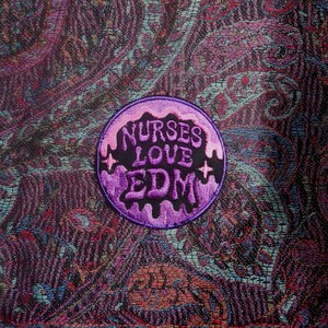 May include: A circular embroidered patch featuring the text NURSES LOVE EDM in a dripping, stylized font. The lettering is purple and black, set against a light purple background with small star accents, all on a dark paisley fabric background.