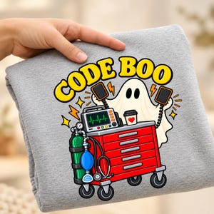 Puede incluir: Una sudadera gris jaspeado con un gráfico de un fantasma blanco sosteniendo paletas de desfibrilador sobre un carro médico rojo. El carro incluye un tanque de oxígeno verde, un resucitador manual azul y un monitor. El texto dice CODE BOO.