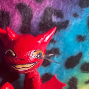 Puede incluir: Una pequeña figura de diablo rojo con orejas puntiagudas, una amplia sonrisa y una cola que termina en forma de corazón. La figura está sentada sobre una base verde redondeada con un acento de rayas moradas. Tiene pequeñas alas rojas brillantes.