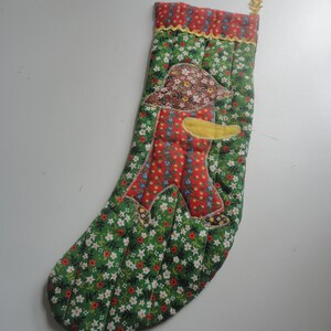 May include: A quilted Christmas stocking with a green floral pattern and a red top. The stocking features an appliqué of a scarecrow with a brown hat and yellow arms. A gold-colored loop is attached to the top.