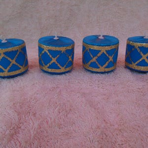 May include: Four cylindrical blue candles arranged in a row. Each candle features a gold glitter lattice pattern wrapping around the exterior side. A small white wick is centered on the flat top surface of every candle in this set of four items.