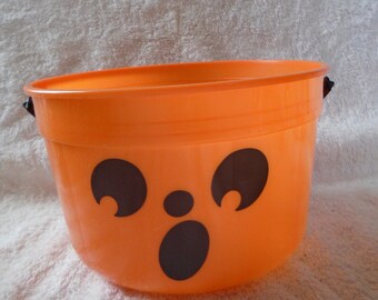 May include: An orange plastic Halloween bucket with a jack-o'-lantern face. The bucket has a wide opening and two small black handles. The face is made of black cutouts for the eyes, nose, and mouth.