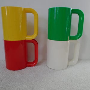 May include: Two stacked mugs, each composed of two colors. One mug is yellow over red, the other is green over white. Both mugs have large, curved handles. The mugs are made of a glossy material.