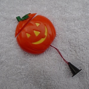 May include: Orange plastic Halloween jack-o'-lantern with a smiling face and a green stem. The pumpkin is split in half, with a red string and a black cone attached. A festive decoration.