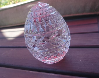 May include: A clear, decorative glass egg with intricate leaf and diamond patterns. The egg-shaped object has a pointed top and is sitting on a dark wooden surface. The glass reflects light, creating a sparkling effect.