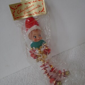May include: Vintage Christmas ornament featuring a doll-like elf with a red hat and green top. The ornament is packaged in a clear plastic bag with a gold tag that reads "Christmas Ornaments". The ornament has a beaded garland.
