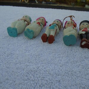 May include: A collection of vintage ceramic figurines, each depicting a small person in various outfits. The figurines have painted details, including facial features, clothing, and accessories. The color palette includes white, blue, red, and black.