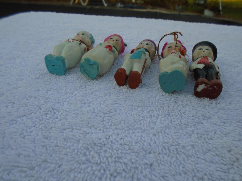 May include: A collection of vintage ceramic figurines, each depicting a small person in various outfits. The figurines have painted details, including facial features, clothing, and accessories. The color palette includes white, blue, red, and black.