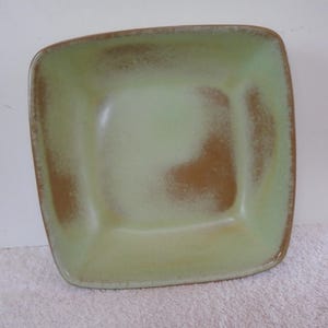 May include: Square ceramic bowl with rounded corners. The bowl has a light green interior with brown accents and a brown rim. The bowl is sitting on a white surface.