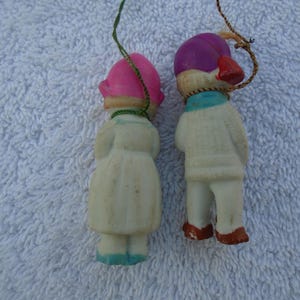 May include: Two vintage Christmas ornaments shaped like children. One wears a pink hat and dress, the other a purple hat and sweater. Both have painted details and hanging strings. The ornaments are made of a cream-colored material.