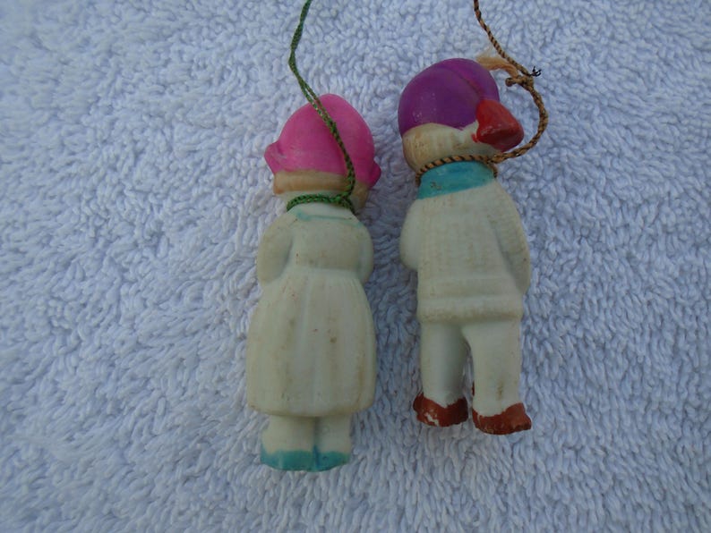 May include: Two vintage Christmas ornaments shaped like children. One wears a pink hat and dress, the other a purple hat and sweater. Both have painted details and hanging strings. The ornaments are made of a cream-colored material.