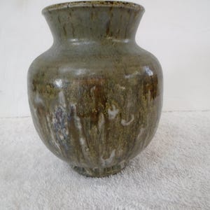 May include: A handcrafted ceramic vase with a wide, rounded body and a flared rim. The vase features a mottled glaze in shades of green, brown, and white, creating a unique, organic appearance. The vase is approximately 8 inches tall.