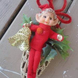 May include: A vintage Christmas ornament featuring a red elf with a smiling face, wearing a matching hat and suit. The elf is nestled in a woven basket with green holly leaves and a gold bell. A red pipe cleaner bow is at the top.