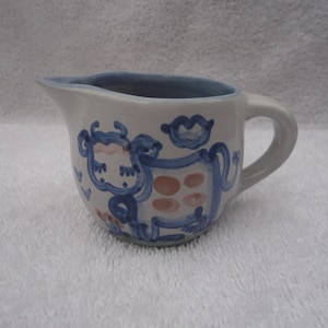 May include: A small, white ceramic creamer with a blue rim and handle. The creamer is decorated with a whimsical blue and orange cow design. The cow has a blue outline and orange spots.