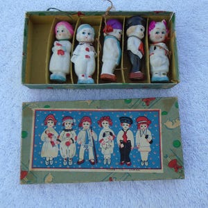 May include: A vintage box set of small porcelain dolls, each dressed in colorful outfits. The dolls are arranged in a divided box, with a matching box below featuring illustrations of similar dolls. The box is labeled "Made in Japan."