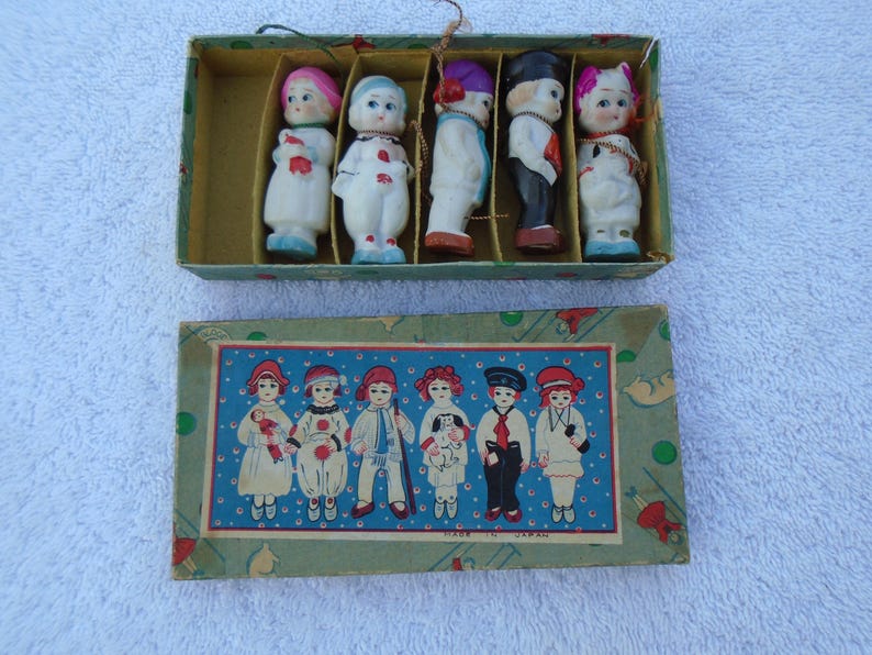 May include: A vintage box set of small porcelain dolls, each dressed in colorful outfits. The dolls are arranged in a divided box, with a matching box below featuring illustrations of similar dolls. The box is labeled "Made in Japan."