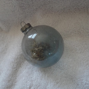 May include: A clear glass Christmas ornament with a silver top and a glittery, frosted finish. The ornament is filled with a dried, brown, plant-like material.