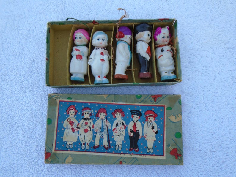 May include: A vintage box set of small porcelain dolls. The dolls are dressed in various outfits with painted faces and hair. The box has a decorative lid with illustrations of similar dolls. The set is marked "Made in Japan".