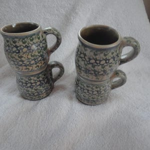 May include: Two ceramic mugs with a speckled green and white glaze. The mugs are stacked on top of each other.