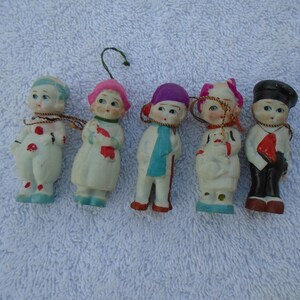 May include: Five vintage porcelain figurines of children, each with unique outfits and expressions. The figurines are painted with details like hats, scarves, and patterned clothing. They have small, painted faces and are arranged on a white surface.