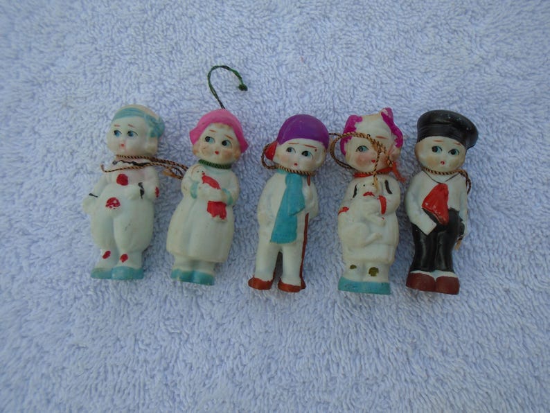 May include: Five vintage porcelain figurines of children, each with unique outfits and expressions. The figurines are painted with details like hats, scarves, and patterned clothing. They have small, painted faces and are arranged on a white surface.
