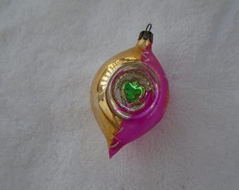 May include: A teardrop-shaped glass Christmas ornament featuring a reflective gold finish on one side and a vibrant hot pink finish on the other. The center contains a circular indentation with a small, shiny green heart-shaped accent inside.