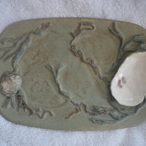 May include: A ceramic serving tray with a green glaze and a raised design of seaweed and oyster shells. The tray has a small, white, ceramic oyster shell in the upper right corner.