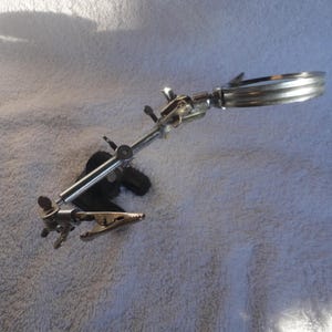 May include: A silver magnifying glass with a clamp base. The magnifying glass is attached to a long, adjustable arm. The arm is attached to a clamp that can be used to secure the magnifying glass to a table or other surface.