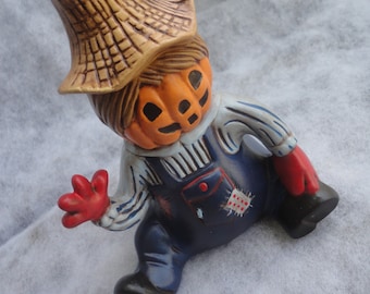 May include: A ceramic figurine of a pumpkin-headed scarecrow wearing blue overalls, a straw hat, and red gloves. The scarecrow has a corn cob on its hat and a stitched patch on its overalls. The figurine is designed for fall or Halloween decor.