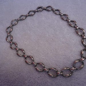 May include: A silver-toned necklace featuring a series of oval-shaped links. The chain is arranged in a circular shape, and the links are connected to each other. The necklace is made of a shiny metal.