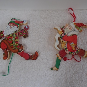 May include: Two flat, jointed wooden Santa Claus ornaments with pull strings. The left Santa wears a red patterned coat and boots. The right Santa wears a red coat with green leggings and holds a white scroll, both featuring red hanging cords.