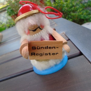 May include: A whimsical wooden figurine of a gnome holding a sign that reads "Sünden-Register". The gnome has a red hat, white beard, and a blue base. The sign is made of wood.