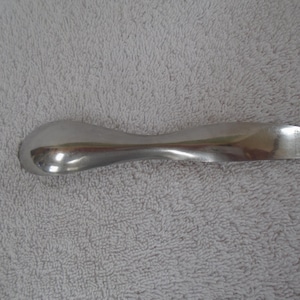 May include: A silver-colored serving spoon with a curved handle and a rounded bowl. The spoon has a rectangular end and is lying on a gray, textured surface.