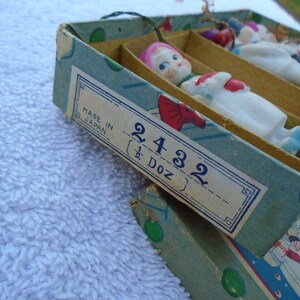 May include: Vintage box of small porcelain dolls, each with a painted face, pink hair, and a white outfit with red and blue accents. The box is labeled "Made in Japan" and "2432 (1/2 Doz)".