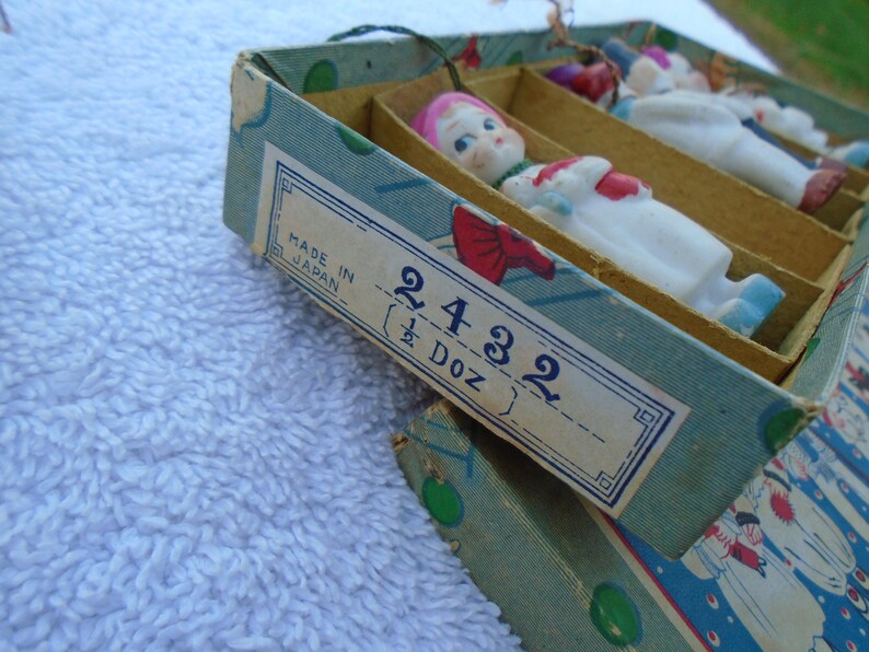 May include: Vintage box of small porcelain dolls, each with a painted face, pink hair, and a white outfit with red and blue accents. The box is labeled "Made in Japan" and "2432 (1/2 Doz)".