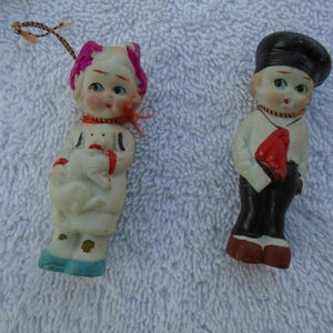 May include: Two vintage porcelain figurines. One is a girl in a white dress holding a dog, with pink accents and blue shoes. The other is a boy in a white shirt, black pants, and a red tie, wearing a black hat.