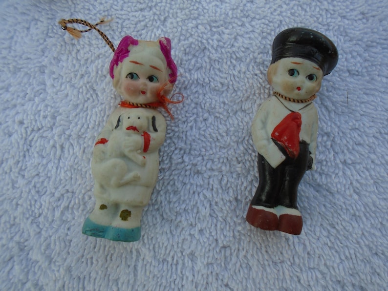 May include: Two vintage porcelain figurines. One is a girl in a white dress holding a dog, with pink accents and blue shoes. The other is a boy in a white shirt, black pants, and a red tie, wearing a black hat.