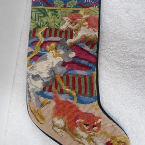 May include: A needlepoint Christmas stocking featuring a blue top border with candy canes and three cats playing with colorful ornaments. The cats are depicted in orange, white, and gray, set against a background of green foliage and red stripes.