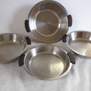 May include: A set of four stainless steel serving dishes. The dishes are round or oval with dark brown handles. The largest dish is round, and the other three are oval. The dishes are on a white surface.