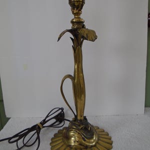 May include: A brass-colored table lamp with a floral design. The lamp features a bulb socket, a decorative stem, and a round base with a scalloped edge. The lamp's cord is visible. The lamp is approximately 15 inches tall.