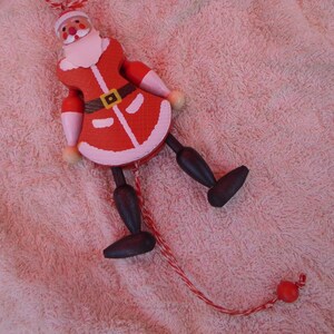 May include: A wooden Santa Claus jumping jack ornament featuring a red coat with white trim, a black belt with a yellow buckle, and dark brown legs. The figure is suspended by a red and white twisted string with a small red bead at the end.