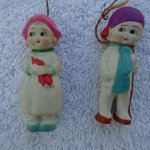 May include: Two vintage porcelain figurines. One is a girl in a white dress, pink hat, and holding a red bird. The other is a boy in a purple hat, blue scarf, and holding a walking stick. Both have painted facial features.