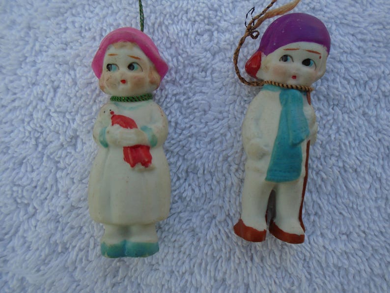 May include: Two vintage porcelain figurines. One is a girl in a white dress, pink hat, and holding a red bird. The other is a boy in a purple hat, blue scarf, and holding a walking stick. Both have painted facial features.