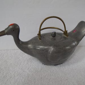 May include: A pewter-colored teapot shaped like a duck, featuring a brass handle and a small red accent on the head. The teapot has a detailed design and a small knob on the lid.